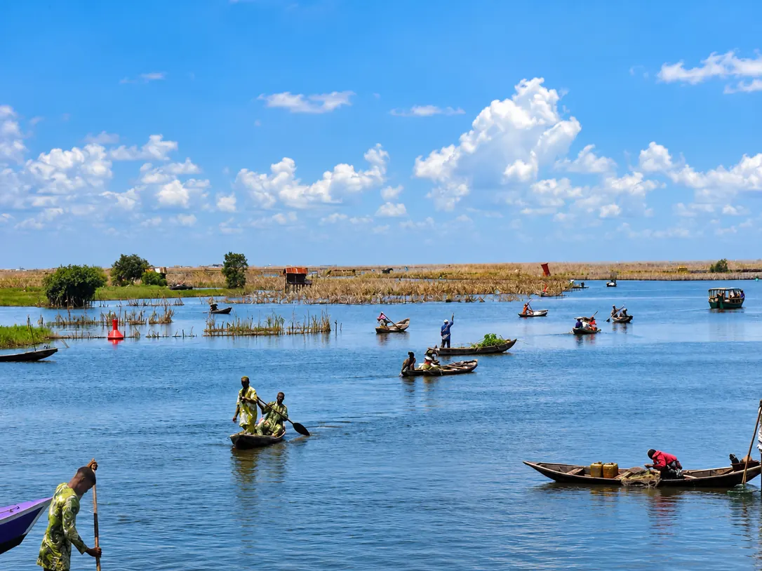 Pirogues et pêcheurs sur un lac sous un ciel bleu, avec des roseaux et une cabane sur pilotis au loin