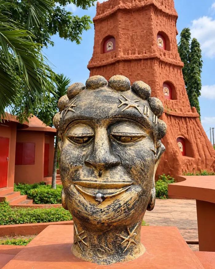 Sculpture d’une tête en bronze devant une tour en terre rouge à Porto-Novo.