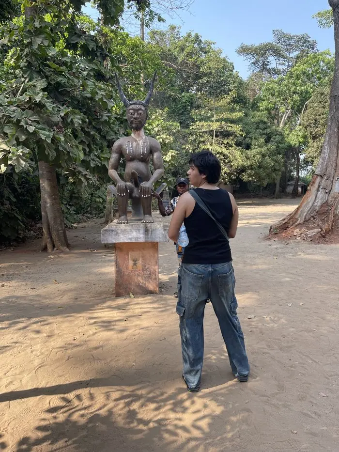 Statue à cornes sur un socle dans une forêt de Ouidah, avec un visiteur et un guide.