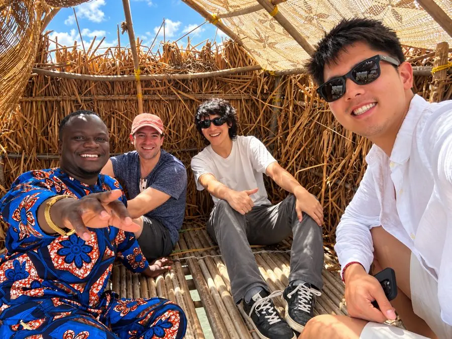 Théophile, guide local à Ganvié, avec trois visiteurs souriants dans une cabane en paille et bambou.