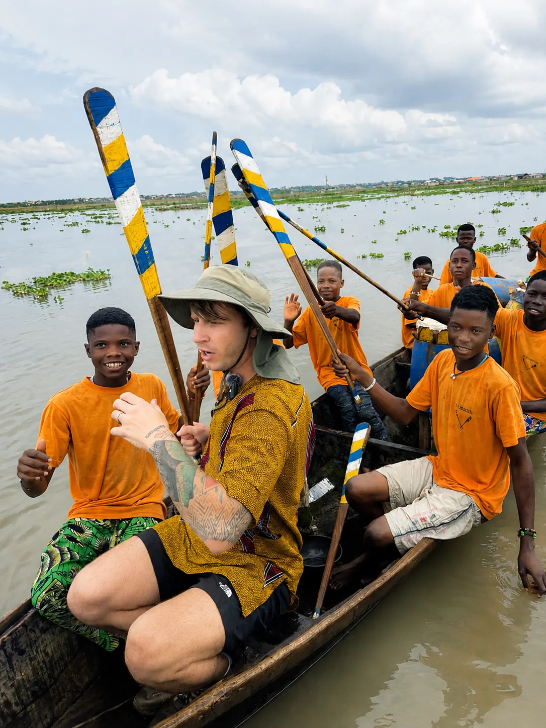 Visiteur assis dans une pirogue avec de jeunes rameurs en maillot orange tenant des pagaies colorées