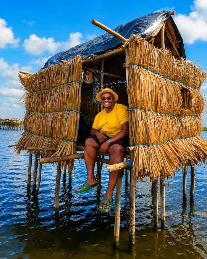 Visiteur souriant assis dans une cabane de paille sur pilotis au-dessus du lac.